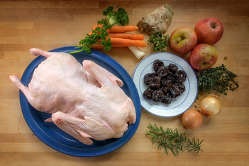 Raw duck ready to cook and ingredients such as apples, baked plums, vegetables and herbs on a wooden kitchen counter, high angle view from above