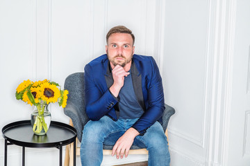 Young American man with beard, relaxing, thinking inside at home in New York City, wearing blue jacket, gray sweater, jeans, sitting on chair, hand touching chin. Yellow Sunflowers on coffee table..