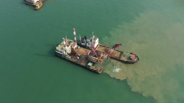 Tilting drone flight of excavating machinery dredging sand and other seabed material, scooping it onto industrial vessel near construction site in Qingdao, China