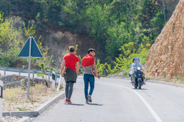 Two male longboarders carrying their longboards in their hands while climbing uphill and preparing for a downhill slide. Wearing red t-shirts, green hat, and super cool sunglasses.