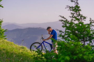 Obraz premium Tired cyclist is wiping his sweat off his face while pushing his bicycle uphill on a dirt road in a mountain.