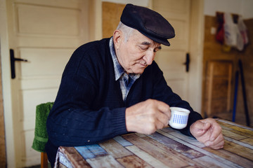 Senior man grandfather old pensioner farmer wearing black sweater and hat having a cup of coffee or tea by the table at home sitting alone
