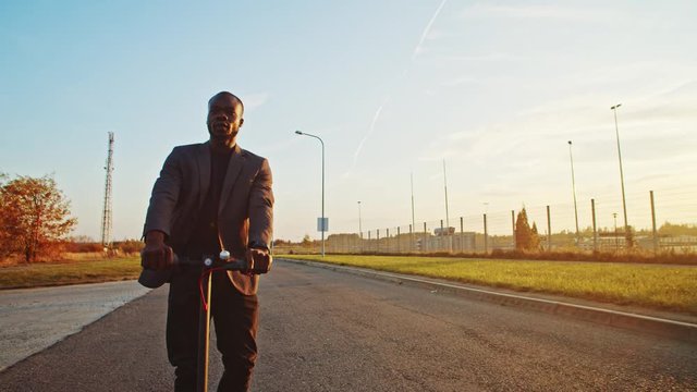 Handsome young african man in business suit riding fast modern e-scooter at sunset. Modern guy traveling in city outskirts by green electric transport.