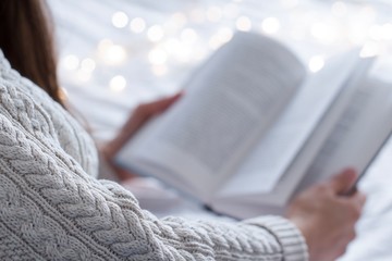 young woman wearing a beige sweater reading a book in a bed with white blanket and gold christmas...