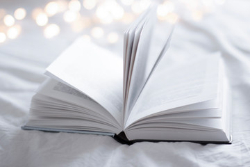 open book on a bed with white blanket with gold christmas lights on background