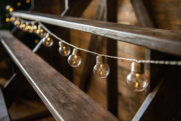 Christmas garland of light bulbs hanging from a wooden staircase. Festive home decoration. Scandinavian loft style. Layout for advertising. Led lamp. Texture background, copy space, blurred banner web