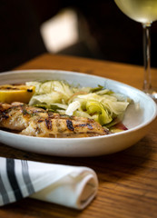 Grilled white fish with lemon and greens on a side with glass of white wine inside restaurant interior