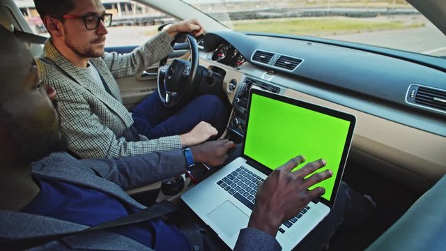 African entrepreneur inside auto using mock-up green chrome key laptop doing car test drive on computer driving with caucasian colleague or friend. Close-up.