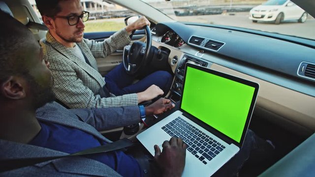 African man driving inside car using laptop with mock-up green monitor close-up. Salesman and customer driving testing car with connected computer device technology.