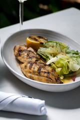 Grilled white fish with lemon and greens on a side with glass of white wine outside restaurant interior