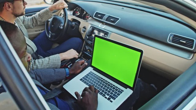 Inside car two diverse successful business partners driving to work. Closeup african man using mock-up laptop green monitor device driving in car.
