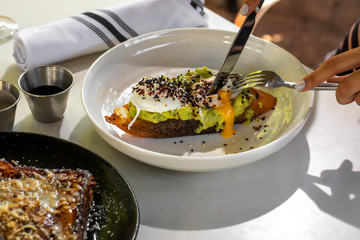 Haute cuisine - grilled sandwich with avocado guacamole sauce and two sunny side up eggs with woman hand and fork outside restaurant in a sunny day