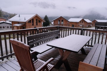 Winter in an alpine chalet.Winter mountain vacation.Wooden houses, table and chairs in the snow.Group of wooden alpine chalets in the mountains in the snow and fog.First snow. Winter season