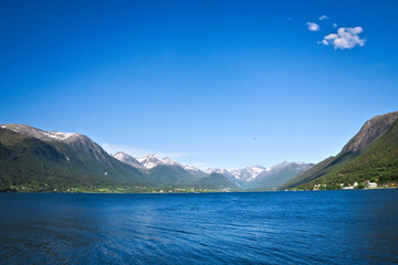 Obraz premium Isfjorden is the beautiful blue fjord at Andalsnes, Norway, with homes at the base of the mountains and snow and clear sky with a couple fluffy clouds in the background on a sunny day.