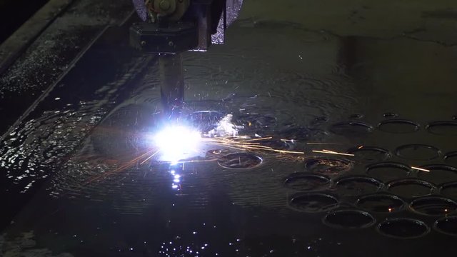 Four times slow-motion shooting of plasma cutting of metal, factory floor, sparks fly, blue flame, huge temperature, water boils, machine makes holes in thick metal sheet, production, heavy industry