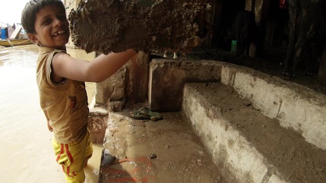 little indian boy shows slipper smeared with cow's shit and washes it in dirty water of holy river Ganges smiles beton stairs hoofs boats Varanasi