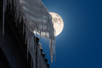 Icicles hang on the roof of a house in the mountains. It is deep in the night and the full moon is partially visible behind the icicles and shines through. © wewi-creative