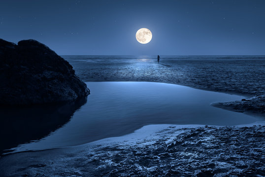 A moon setting on the beach in Cornwall in the early morning. It's low tide and the moonlight is reflected in the water. A woman goes towards the moon.
