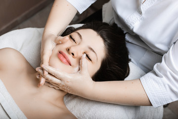Upper view portrait of a lovely young woman having facial massage by a cosmetologist in a spa center.