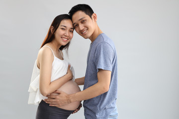 young asian husband standing and embracing young asian pregnant belly wife with happiness and smiling face on white background and looking at camera