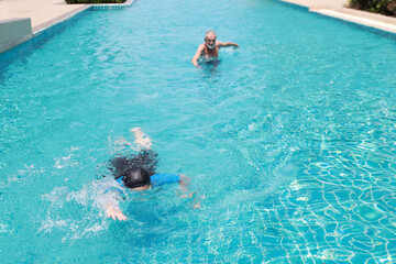 high angle view happy elderly caucasian husband and elderly asian wife swimming in pool during retirement holiday with relaxation on sunny day