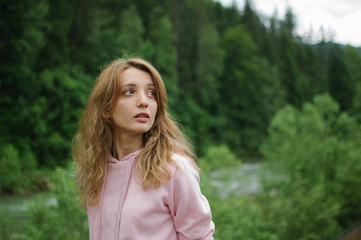 Amazing young girl in pink casual clothes with long hair on the hill with green forest background during spring or early autumn in the mountains.