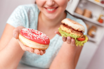 Junk Food. Chubby girl sitting at kitchen choosing between hamburger and doughnut close-up smiling joyful blurred