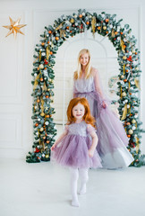 Redhead daughter and her blonde mother are playing before arch of fir branches in a Christmas studio