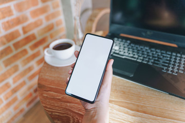 Woman holding smartphone with blank white screen on wooden table at coffee shop.Blank screen mobile phone with coffee cup.