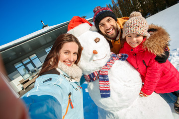 Winter vacation. Family time together outdoors standing near house taking selfie with snowman...