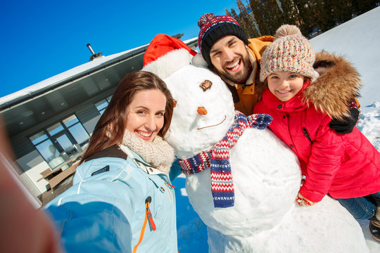 Winter vacation. Family time together outdoors standing near house taking selfie with snowman smiling happy close-up