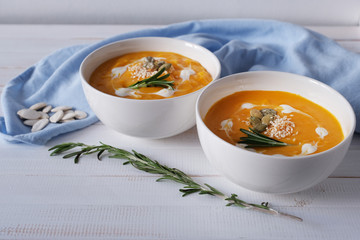 Two bowls of pumpkin soup with rosemary, seeds and cream on white wood background