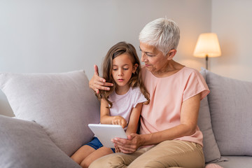 Smiling granddaughter and grandmother using digital tablet on sofa in living room. A grandmother...