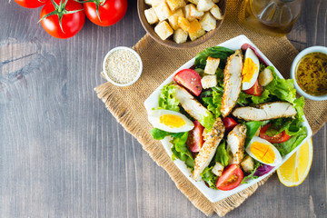 Fresh Caesar salad with delicious chicken breast, ruccola, spinach, cabbage, arugula, egg, parmesan and cherry tomato on wooden background. Oil, salt and pepper. Healthy and diet food concept.