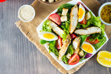 Fresh Caesar salad with delicious chicken breast, ruccola, spinach, cabbage, arugula, egg, parmesan and cherry tomato on wooden background. Oil, salt and pepper. Healthy and diet food concept.