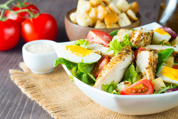 Fresh Caesar salad with delicious chicken breast, ruccola, spinach, cabbage, arugula, egg, parmesan and cherry tomato on wooden background. Oil, salt and pepper. Healthy and diet food concept.