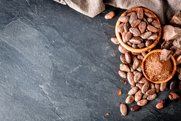 Cocoa beans on dak stone table top view. Dark chocolate and powder in wooden bowl copy space for text or banner.