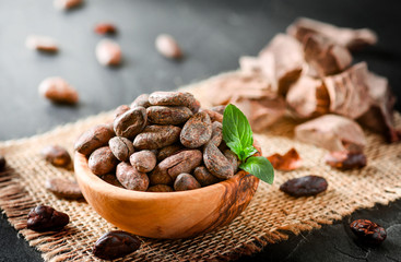 Cocoa beans with mint on jute side view. Cacao powder in wooden bowl blure in background. Panorama or banner concept.