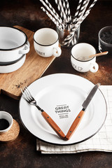 Crockery and cutlery on a dark background with copy space. White metal plates, bowls, cups and cutlery with wooden handles.