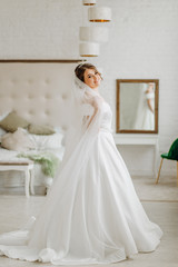Fototapeta premium Pretty bride in a white dress stands in a room by the bed