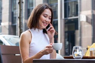 Smiling Asian woman drinking coffee and using her mobile phone. Satisfied female enjoying cup of coffee. Close up portrait of beautiful girl drinking coffee from a white mug in the coffee shop