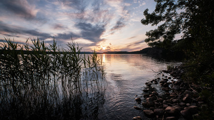 Sunset over a lake with rocks and reeds in the foreground and the sun in the background