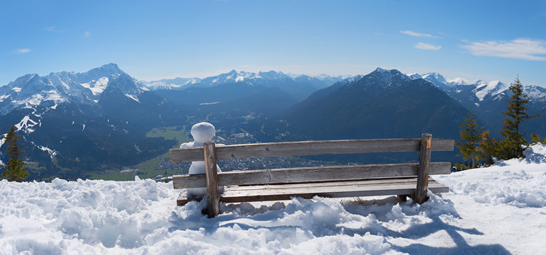 kleiner Schneemann sitzt auf einer Bank mit Blick nach Garmisch und die Zugspitze, Winterlandschaft
