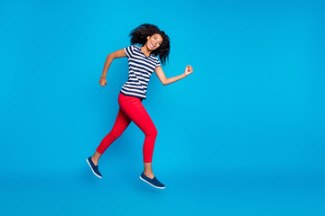 Fototapeta na wymiar Full length profile side photo of cheerful crazy afro american girl jump run fast after spring discounts wear striped t-shirt red pants trousers sneakers isolated over blue color background