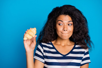 Close up photo of addicted funky afro american girl hold croissant look bite lips want eat think its yummy tasty pastry food wear stylish striped t-shirt isolated over blue color background