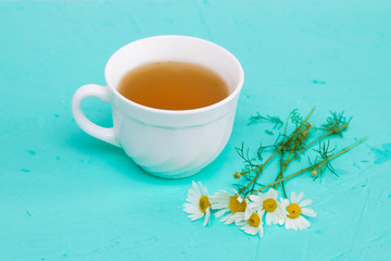 Chamomile tea in a white tea cup, chamomile flowers on the table, turquoise background.