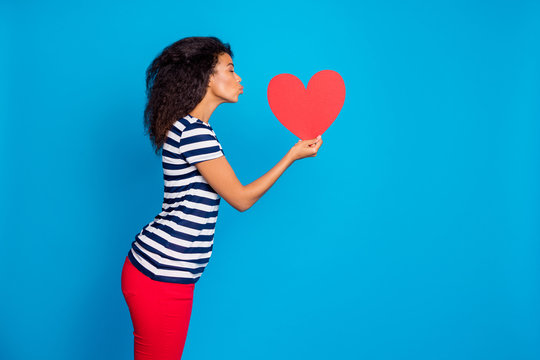 Side profile photo of tender soft cute adorable girlfriend kissing big red heart inspired by her love near empty space wearing striped t-shirt isolated vivid color blue background