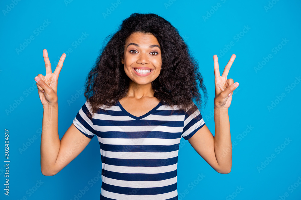 Wall mural Photo of cheerful positive nice pretty cute charming girlfriend showing you double v-sign showing her white teeth in striped t-shirt isolated vivid color blue background