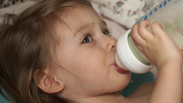 Сlose up of little cute baby girl lies on bed after sleep in morning and sucks milk from bottle with great appetite. Baby food. Infant formula milk concept