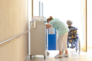 Aide soignante au travail accompagne personne agée en fauteuil roulant dans couloir hopital clinique maison de retraite - obrazy, fototapety, plakaty